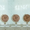 A photograph of three 2021 Gruber Cosmology Prize trophies set on a concrete surface. The trophy primarily consists of a gold coin on a semi-meridian mount similar to the one found on an Earth globe model, so that the coin can rotate in place. The coin and its mount are attached to a rectangular glass base. The coin is engraved with an icon of a rocket in the midst of takeoff, a simplified icon of the Milky Way, Einstein's equation of 