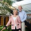 A photograph of Marilyn and Jim Simons. Marilyn is a woman with light blonde hair and is wearing a pink, black, and white button sweater with a white undershirt and black slacks. Jim Simons is a man with a faint white beard, a blue-and-white pinstripe button-down shirt, and beige slacks. They are situated in a room with a staircase leading upward and that has a small garden with a tree underneath it.