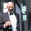 A photograph of David Spergel, a bald bearded man with yellow circular glasses, in a black suit with a white shirt and black bowtie. He is situated in an outdoor setting where there are people similarly dressed to him standing in the background. He is smiling at the camera and is holding a flute of champagne.