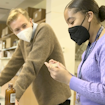 A photograph of two people wearing masks and working at a desk in a laboratory-like setting. One of the people is holding a small container in their hand.